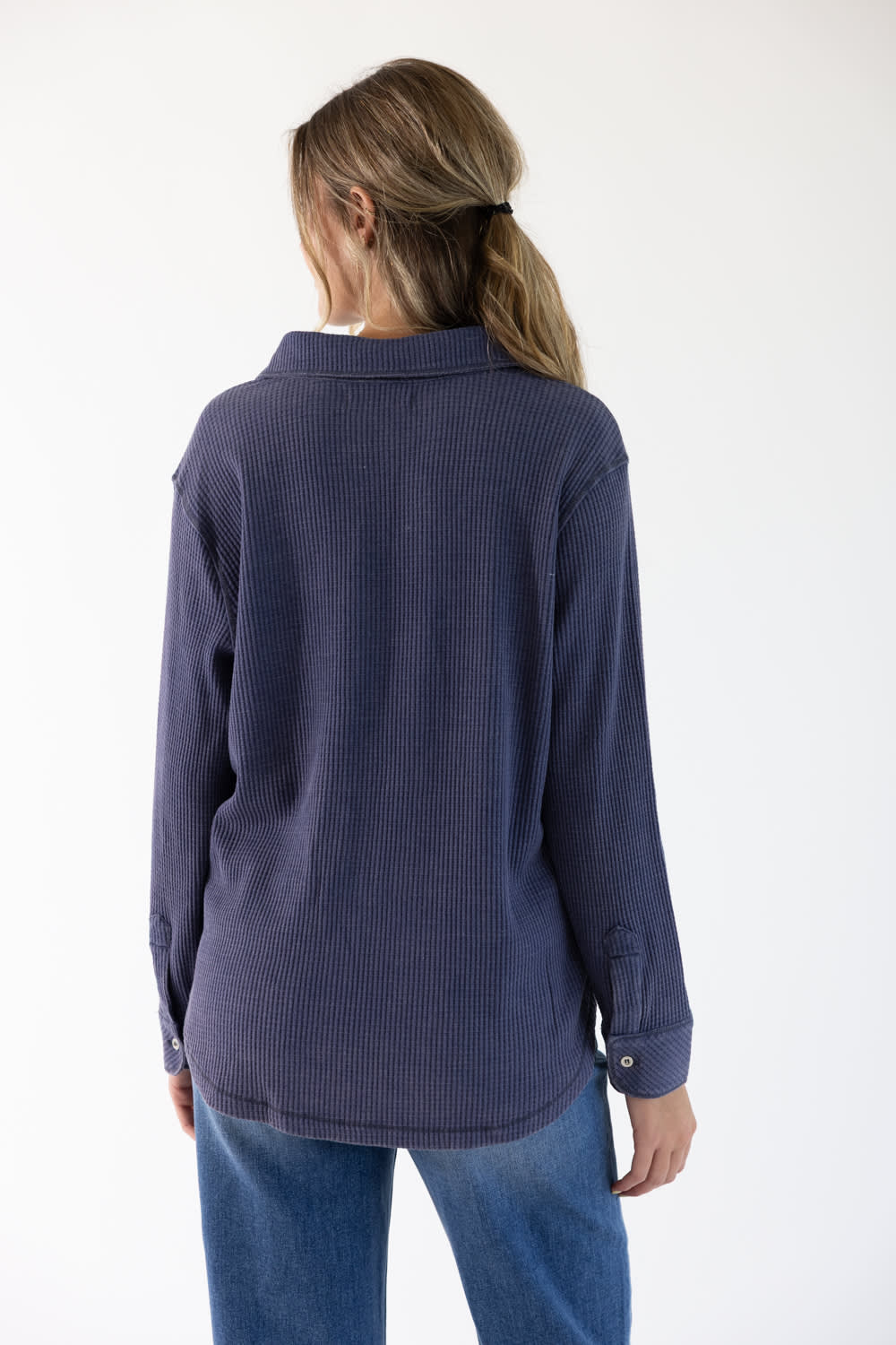 A woman viewed from the back wearing a dark navy blue ribbed long-sleeve shirt and blue jeans against a plain white background.