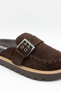 A close-up of a dark brown suede mule slide featuring a decorative strap with a silver-tone metal buckle, decorative stitching, and a thick dark brown lug sole.