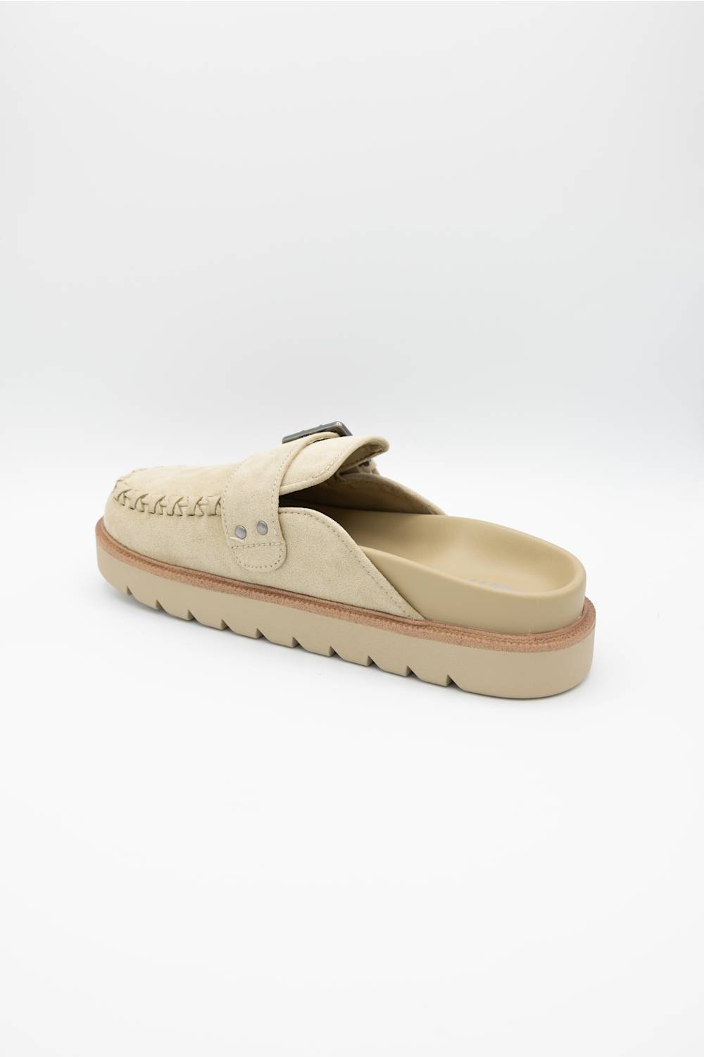 A single beige slip-on clog-style shoe with a textured sole, decorative stitching, and a small metal buckle detail, shown from a side angle on a white background.