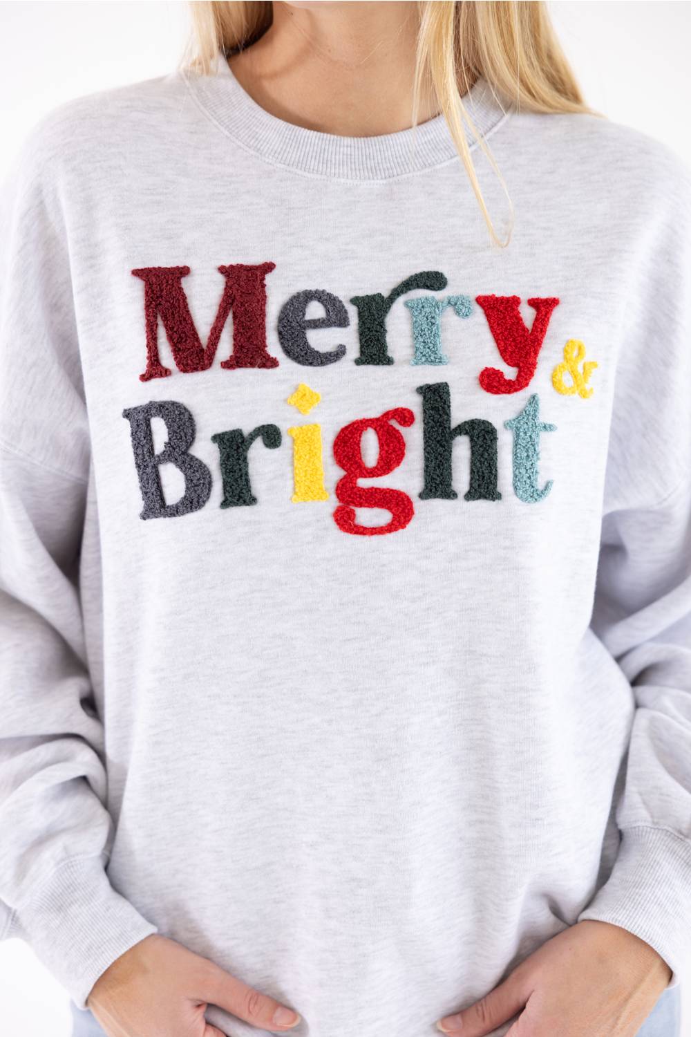 A person wearing a light heather gray crewneck sweatshirt with the words 'Merry & Bright' printed across the chest in multicolored textured letters.
