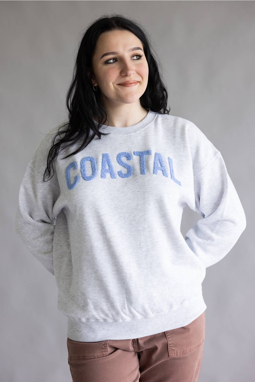 A woman wearing a light heather gray crewneck sweatshirt with 'COASTAL' embroidered in blue across the chest, paired with dusty rose pants, against a neutral gray background.