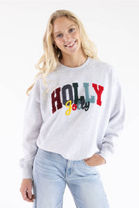 A woman wearing a light heather gray sweatshirt with a colorful 'HOLLY Jolly' graphic on the front, paired with light blue jeans.