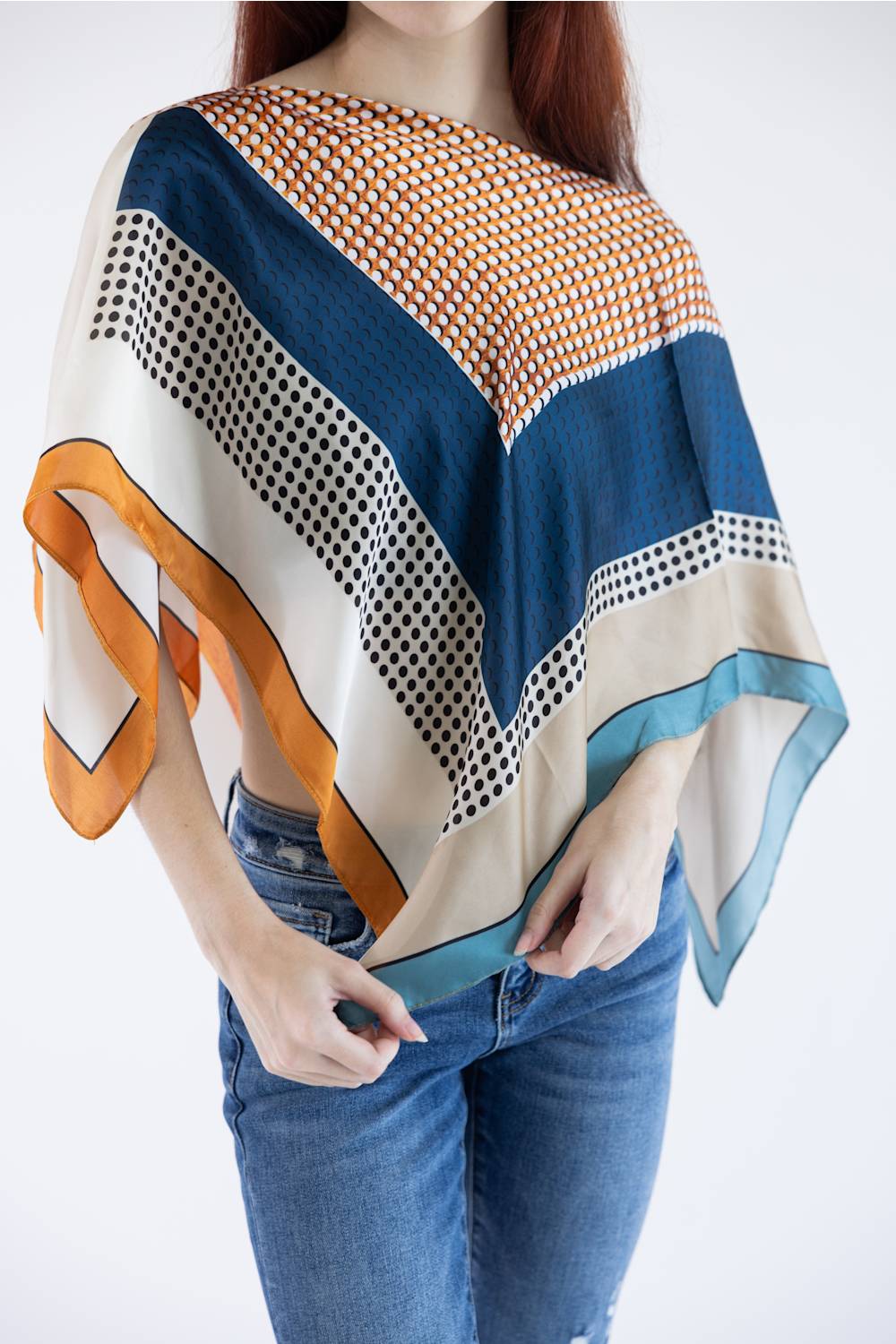 A woman wearing a loose, wide-sleeved patterned shawl or poncho in blue, cream, orange, and black over blue jeans, photographed against a plain white studio background.