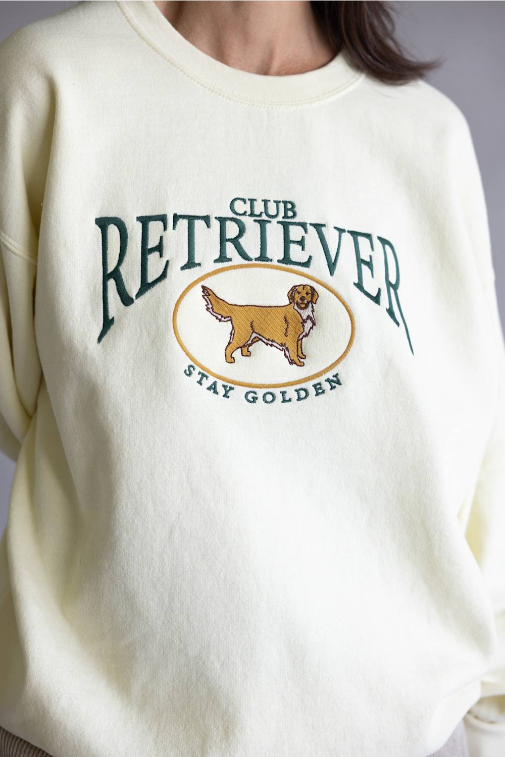A close-up of a cream-colored crewneck sweatshirt featuring a green and gold graphic that reads 'CLUB RETRIEVER' and 'STAY GOLDEN' with an illustration of a golden retriever dog.