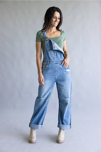 A woman modeling light-wash blue denim overalls with a green short-sleeve top and beige ankle boots, standing against a plain light gray studio background.