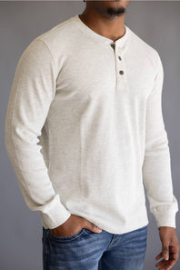 A man wearing a light cream/off-white long-sleeve henley shirt with a three-button placket, paired with blue jeans, shown from the waist up against a neutral gray background.