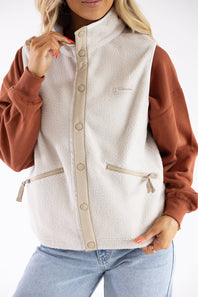 A woman wearing a cream sherpa fleece vest with tan trim over a rust-brown long-sleeve top and light blue jeans, shown from the neck down against a white studio background.