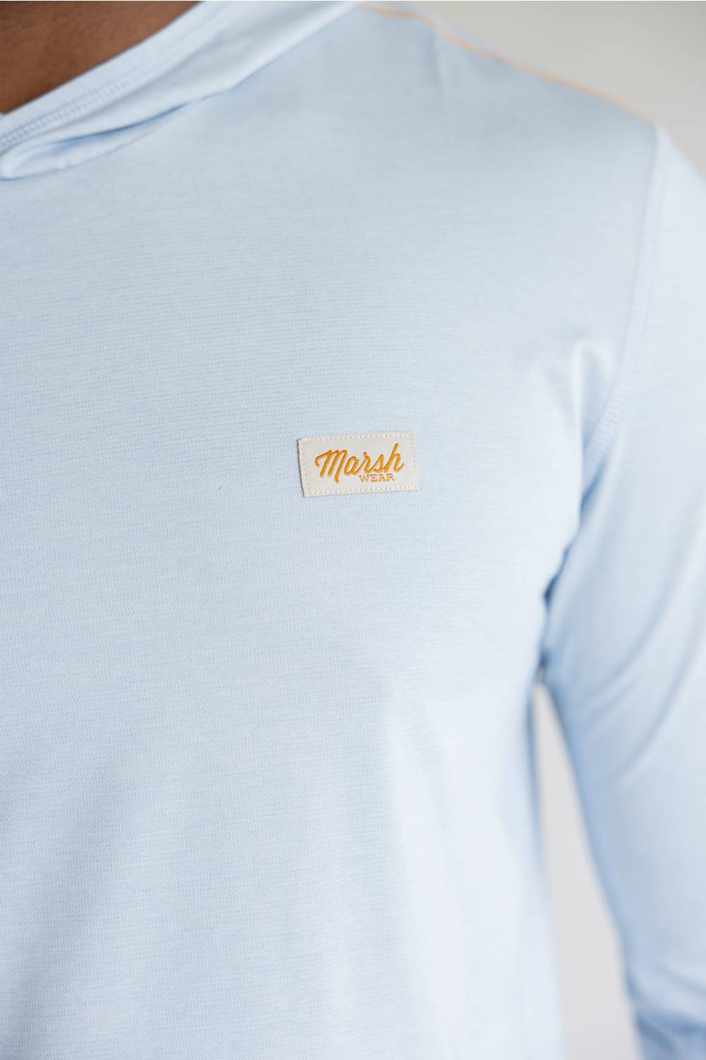 A close-up of a light blue long-sleeve t-shirt worn by a person, featuring a small rectangular tan patch on the left chest with the brand name 'Marsh' in orange script.