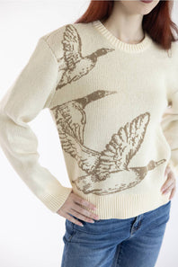 A cream-colored knit sweater featuring a brown graphic print of three flying ducks, worn by a model with blue jeans against a white background.
