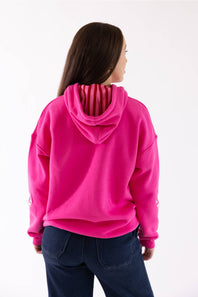 A woman viewed from the back wearing a bright pink pullover hoodie with dark blue jeans, set against a plain white studio background.