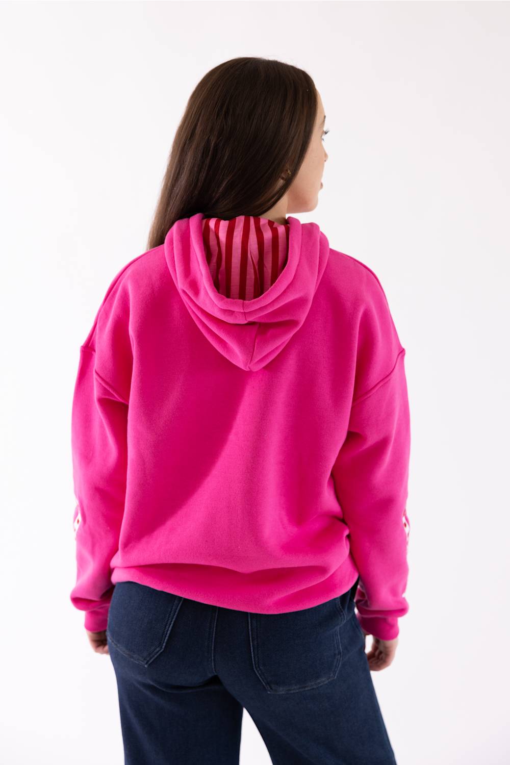 A woman viewed from the back wearing a bright pink pullover hoodie with dark blue jeans, set against a plain white studio background.