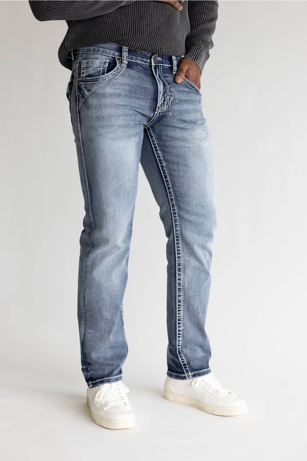 A person wearing light-wash blue jeans with a dark gray ribbed sweater and white sneakers, standing against a plain light gray studio background.