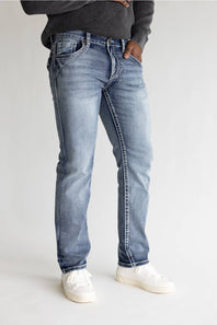 A person wearing light-wash blue jeans with a dark gray ribbed sweater and white sneakers, standing against a plain light gray studio background.