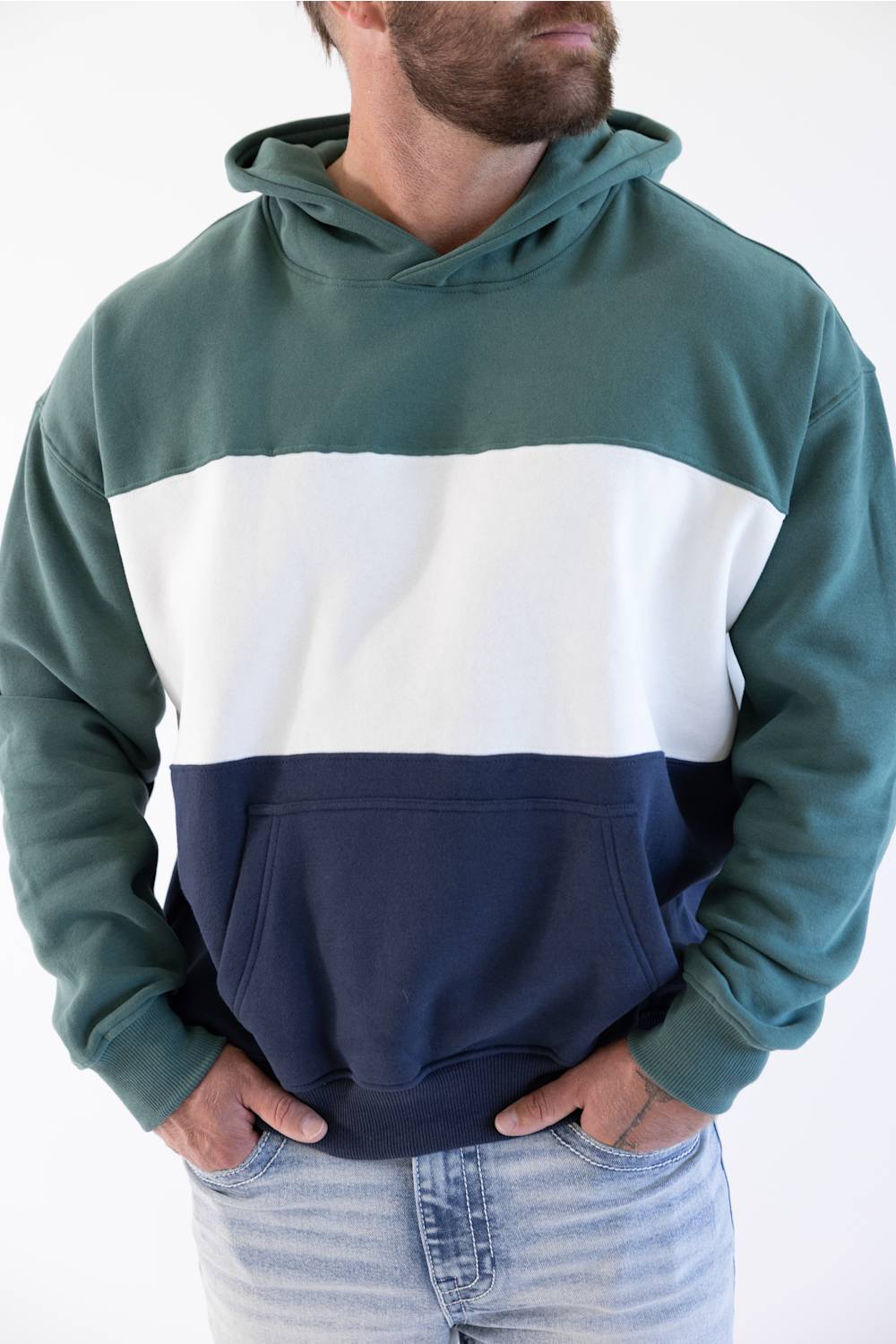 A man wearing a color-block hoodie with a dark green top, a wide white middle stripe, and a navy blue bottom, paired with light-wash jeans against a plain white background.