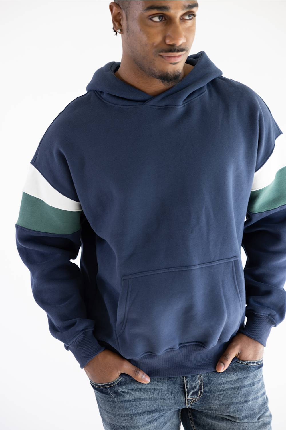 A man wearing a navy blue pullover hoodie with color-blocked sleeves in white and green, paired with blue jeans, against a plain white background.