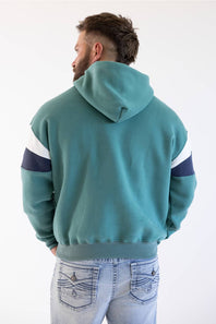 A man viewed from behind wearing a teal colorblock hoodie with white and navy sleeve stripes, paired with light-wash blue jeans, against a plain white background.