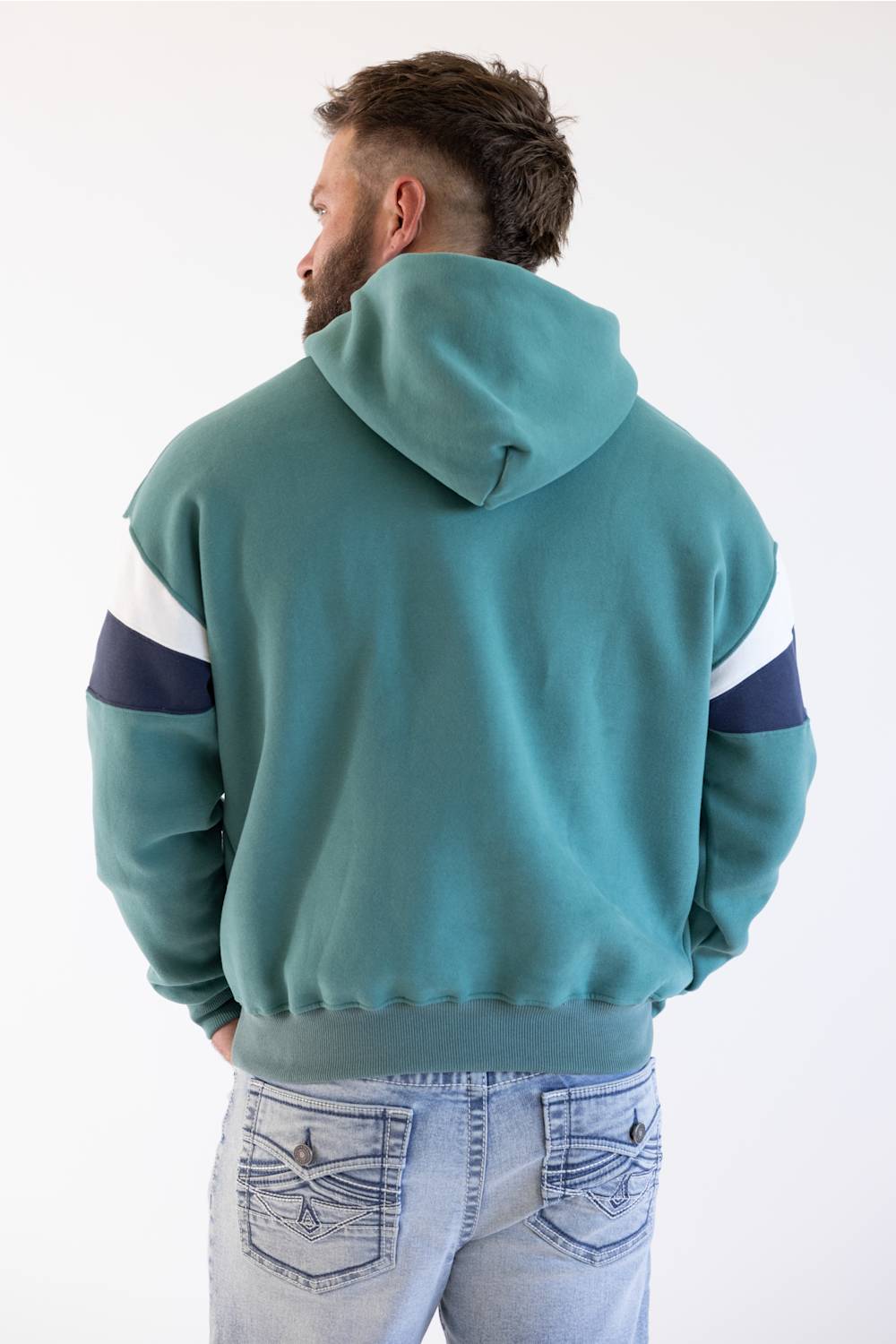A man viewed from behind wearing a teal colorblock hoodie with white and navy sleeve stripes, paired with light-wash blue jeans, against a plain white background.