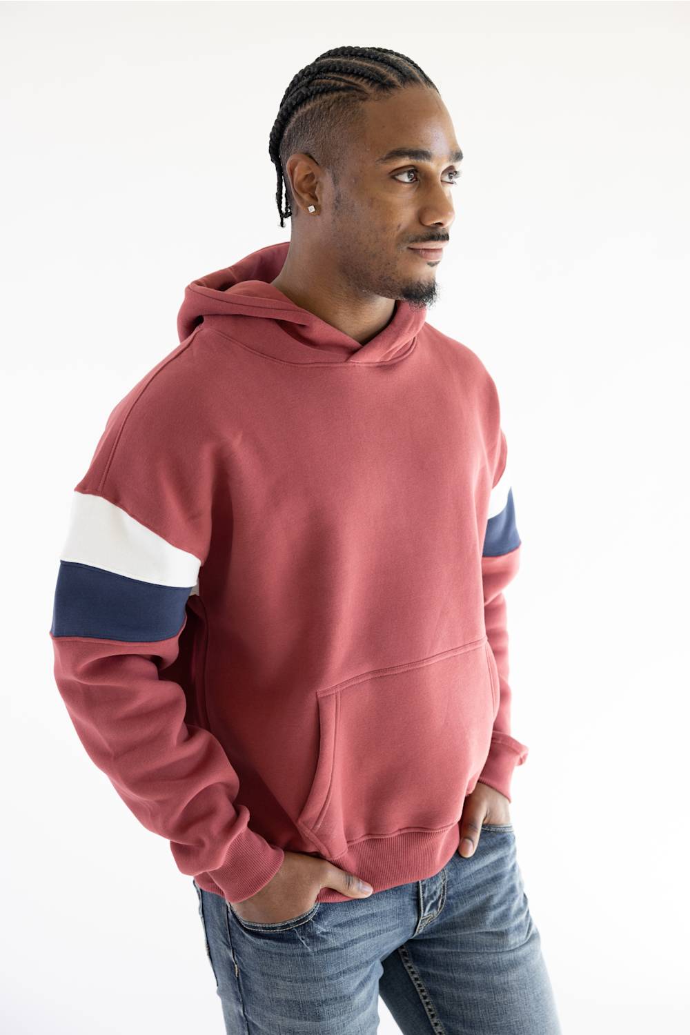 A man wearing a dusty rose pink colorblock hoodie with navy and white stripes on the sleeves, paired with blue jeans, photographed against a clean white background.