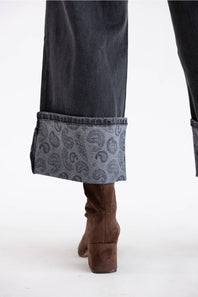 Close-up of the lower legs of a person wearing dark gray wide-leg jeans with a folded cuff showing a paisley pattern, paired with brown suede ankle boots, against a plain white studio background.