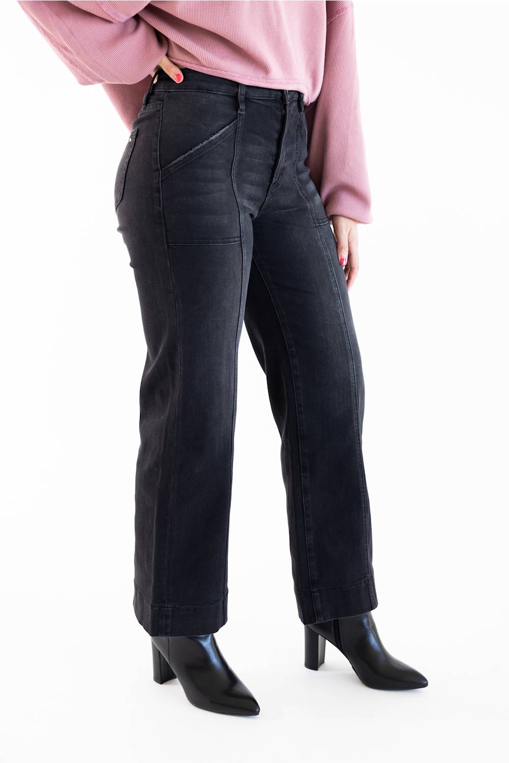 A model wearing a pair of dark washed black wide-leg jeans, styled with a dusty pink long-sleeve top and black pointed-toe heeled boots, shown from the waist down on a white studio background.