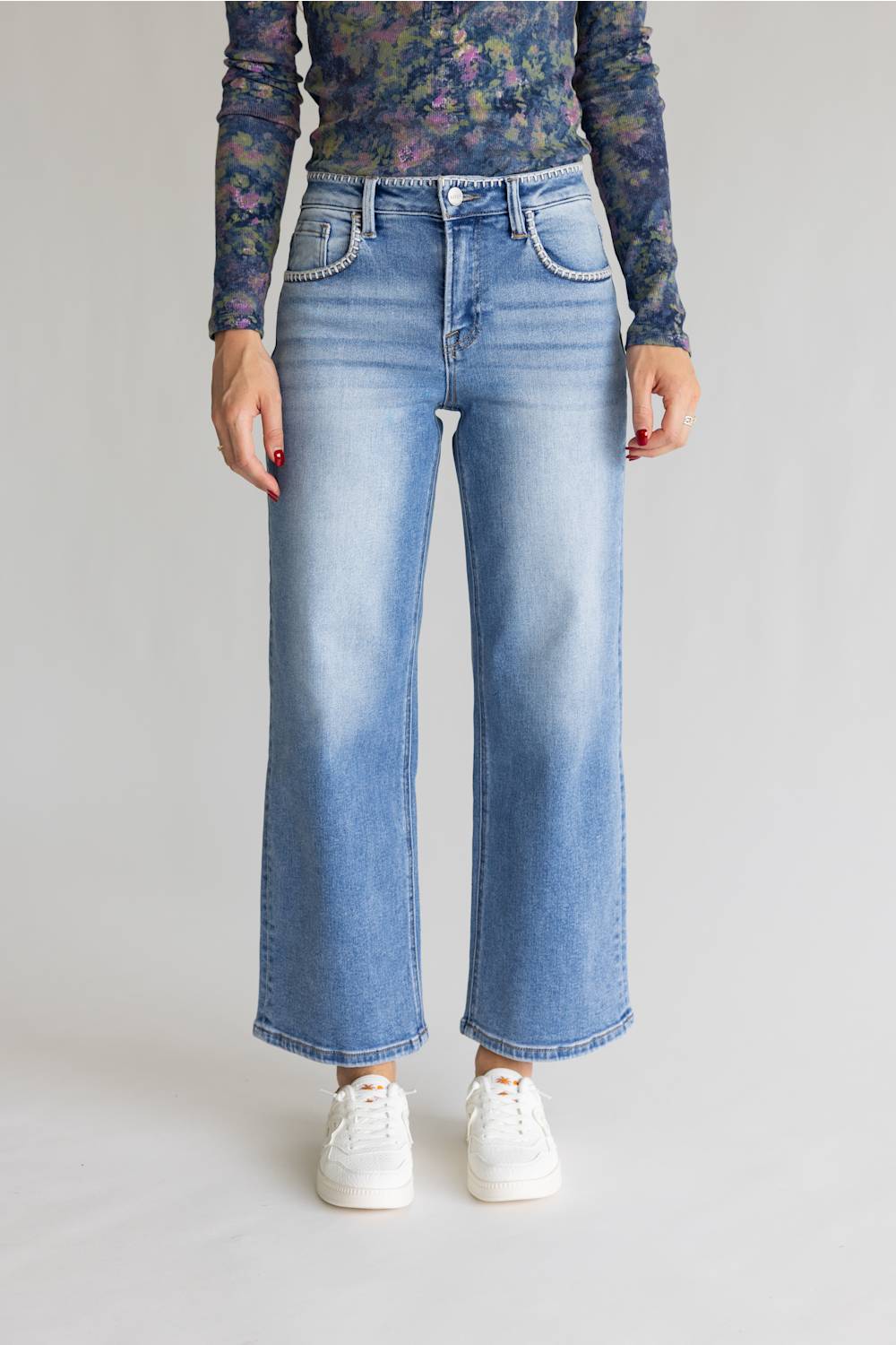 A model is shown from the waist down wearing light blue wide-leg denim jeans, a dark floral long-sleeve top, and white sneakers against a neutral studio background.