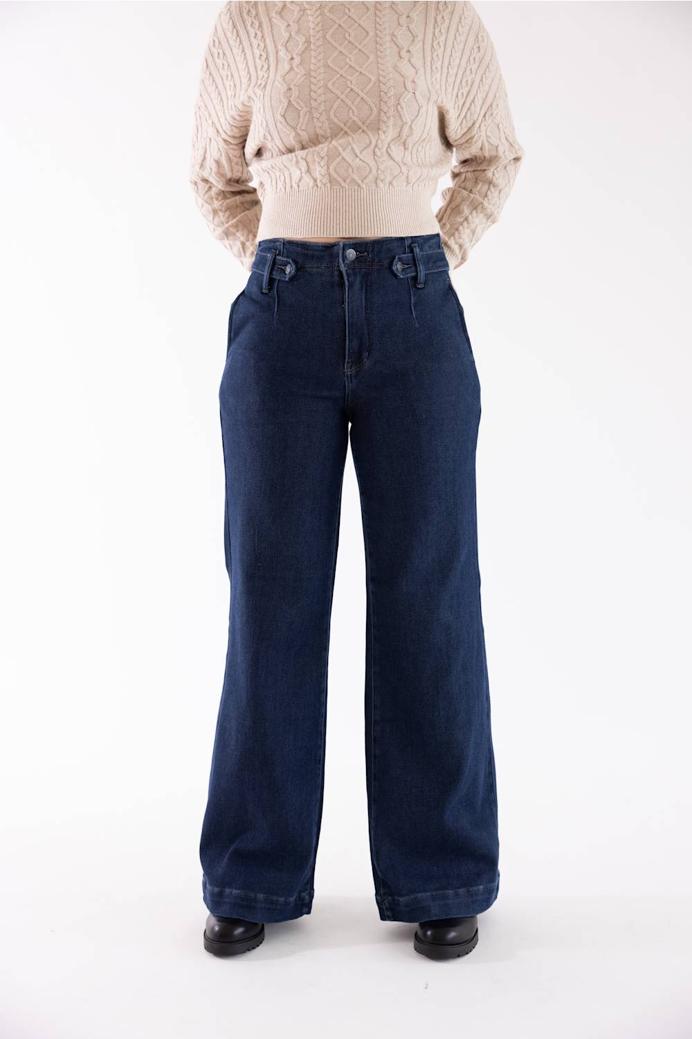 A person wearing dark blue wide-leg denim jeans and a beige cable-knit sweater, standing against a plain white studio background.
