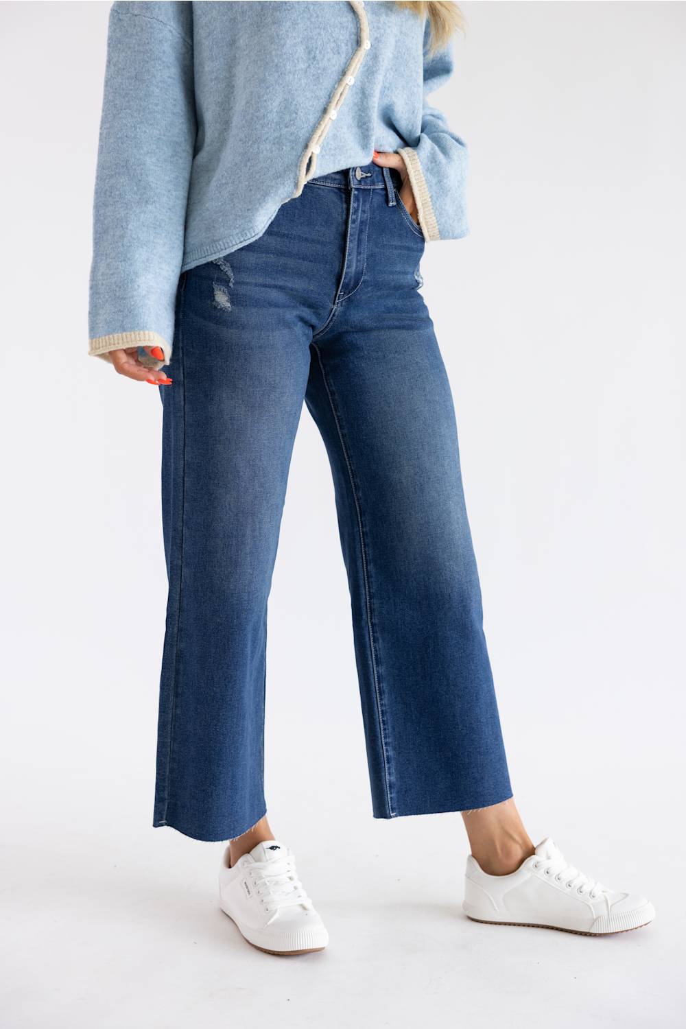 A person wearing blue cropped wide-leg jeans with a light blue knit top and white sneakers, photographed against a clean white studio background.