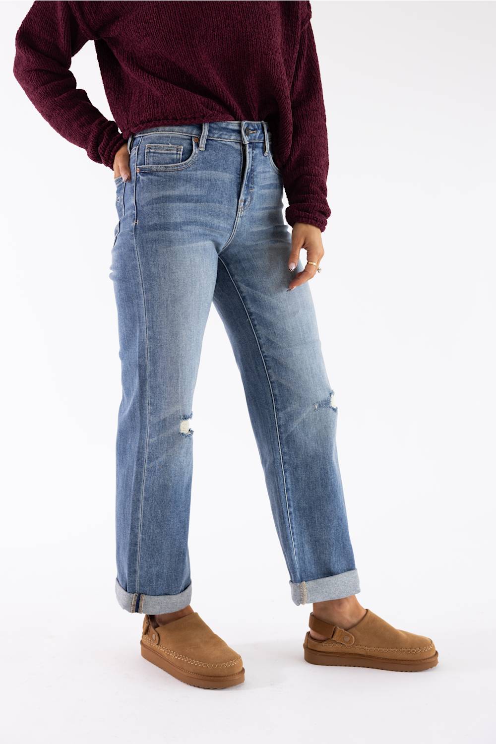 A person wearing light-wash blue distressed jeans with rolled cuffs, paired with a dark burgundy sweater and brown slip-on shoes, shown from the chest down on a plain white studio background.
