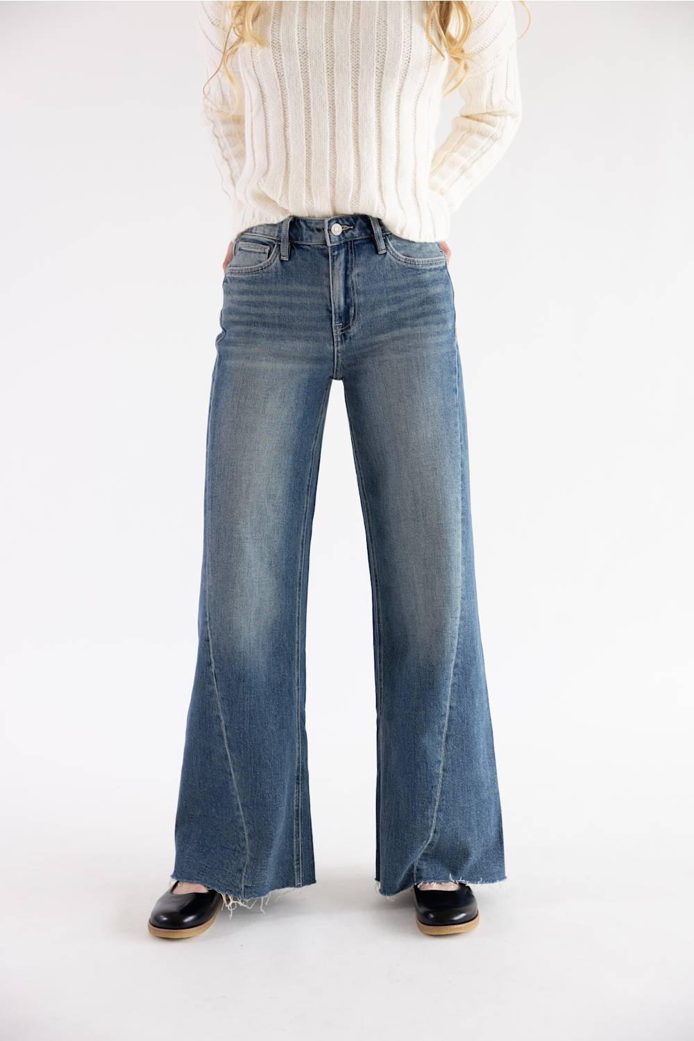 A model wearing a pair of high-waisted, wide-leg blue denim jeans with a faded wash, styled with a cream ribbed knit sweater and black shoes against a plain white studio background.