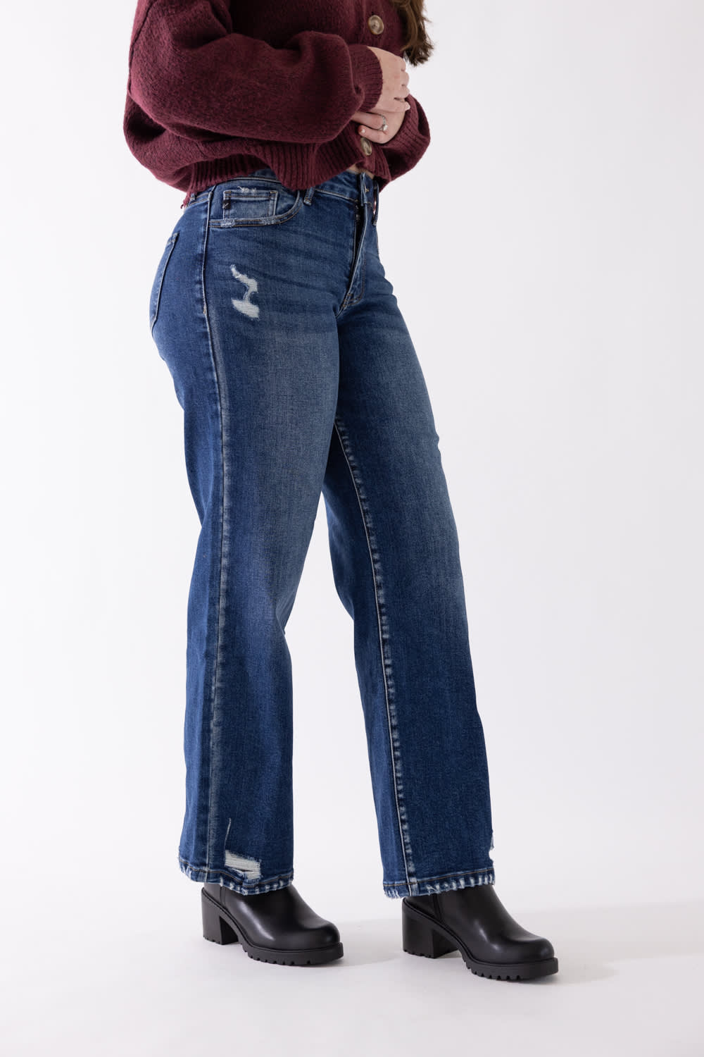 Side view of a model wearing KanCan dark wash, high-rise wide-leg jeans with subtle distressing near the knee and hem, styled with black boots and a burgundy sweater against a plain backdrop