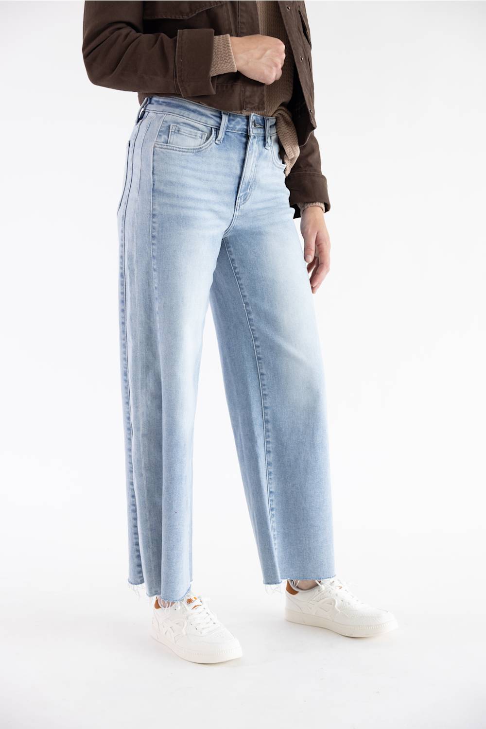 A model wearing light-wash wide-leg denim jeans, styled with a brown jacket and white sneakers, photographed against a seamless white studio background.