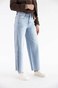 A model wearing light-wash wide-leg denim jeans, styled with a brown jacket and white sneakers, photographed against a seamless white studio background.