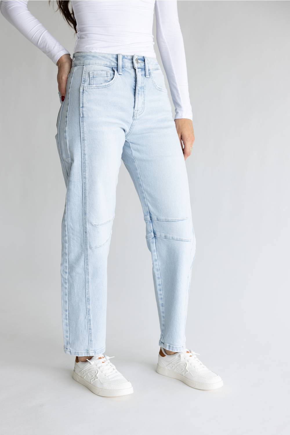 A model wearing light-wash light blue denim jeans with distinctive seam detailing running down the sides of the legs, styled with a white long-sleeve top and white sneakers against a light gray studio background.