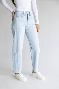 A model wearing light-wash light blue denim jeans with distinctive seam detailing running down the sides of the legs, styled with a white long-sleeve top and white sneakers against a light gray studio background.