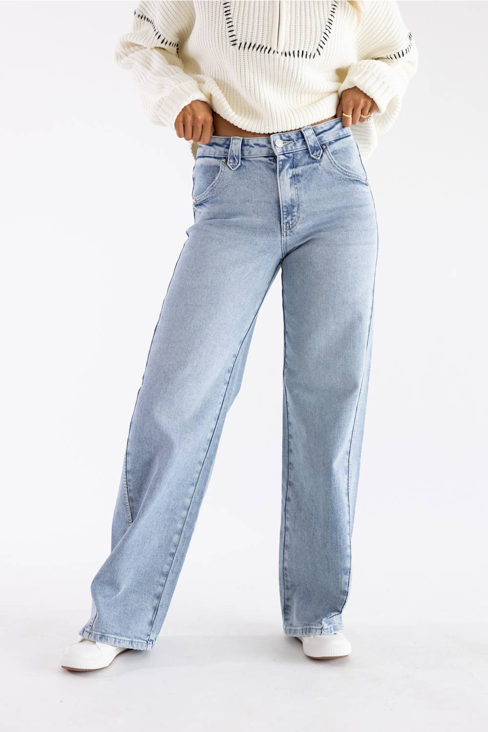 A person wearing light-wash wide-leg blue jeans, a cream knit sweater, and white pointed shoes, standing against a plain white studio background.