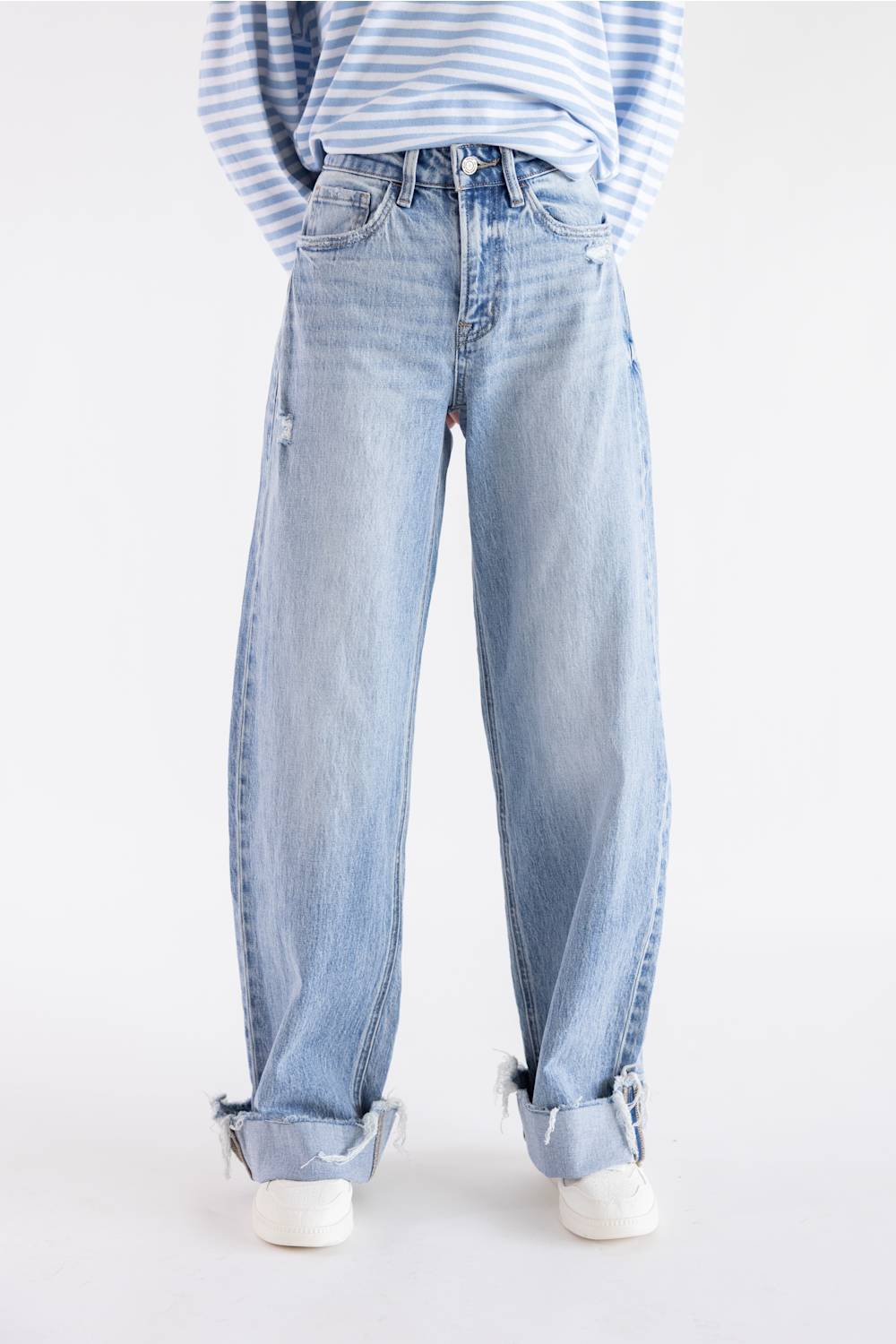 A person wearing light-wash blue wide-leg jeans with rolled cuffs, paired with a blue-and-white striped long-sleeve top and white sneakers, photographed against a plain white studio background.