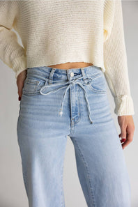 A person wearing light-wash blue jeans with a drawstring waist and a cream ribbed knit sweater, shown from the chest down against a plain light gray studio background.