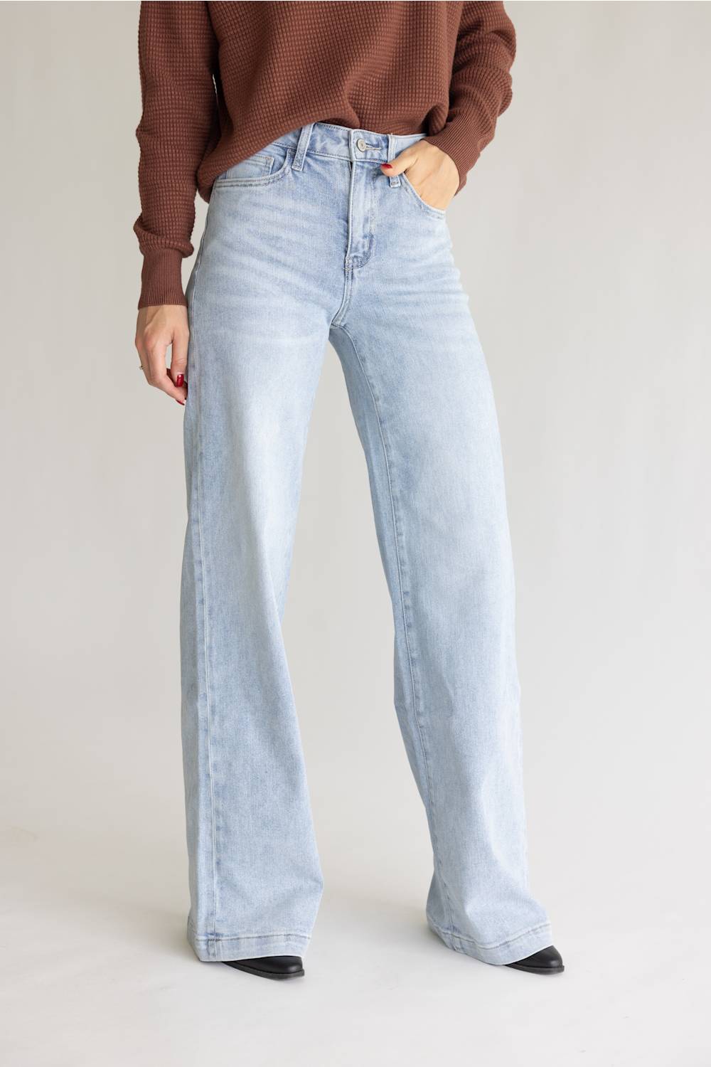 A model wearing light-wash, wide-leg denim jeans and a brown ribbed knit sweater, standing against a neutral light gray studio background.