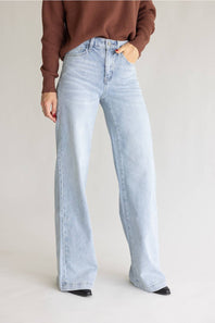 A model wearing light-wash, wide-leg denim jeans and a brown ribbed knit sweater, standing against a neutral light gray studio background.
