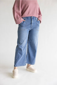 A person wearing light blue wide-leg cropped jeans, a dusty pink long-sleeve top, and cream slip-on shoes, standing against a plain light gray studio background.