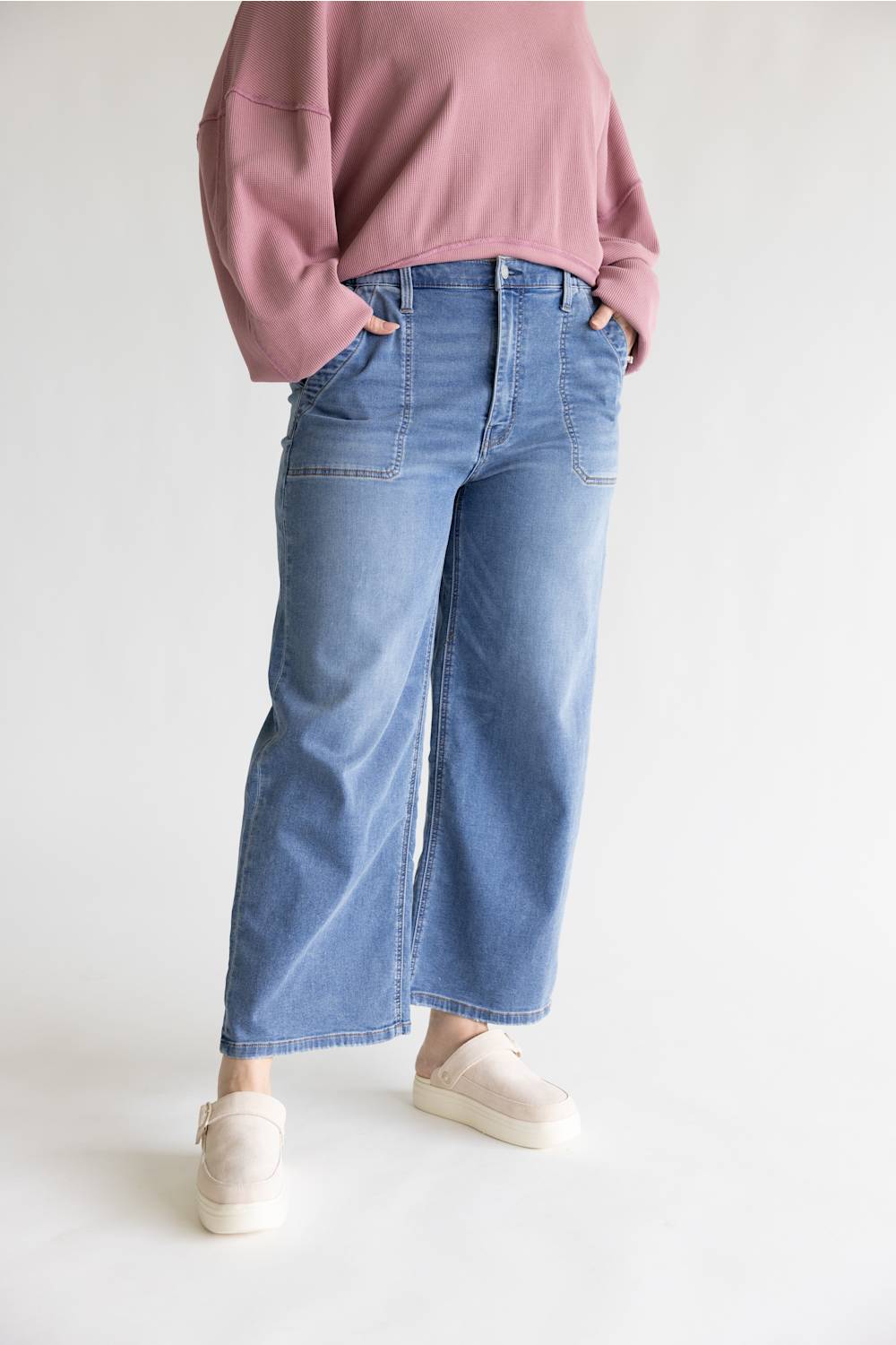 A person wearing light blue wide-leg cropped jeans, a dusty pink long-sleeve top, and cream slip-on shoes, standing against a plain light gray studio background.