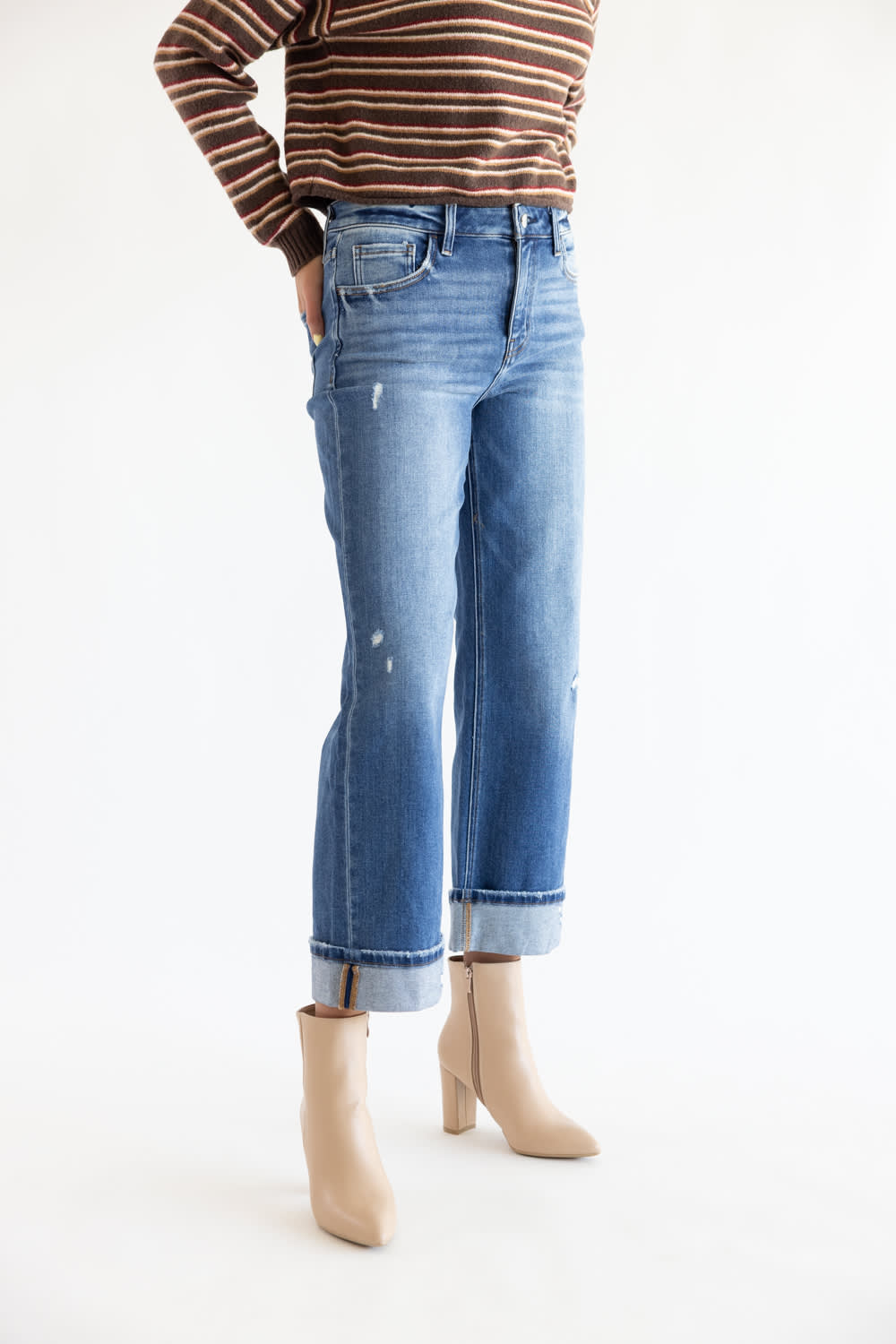 A person wearing light-wash blue cropped jeans with rolled cuffs, paired with a brown and beige striped long-sleeve top and beige ankle boots, photographed against a plain white studio background.