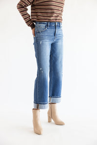 A person wearing light-wash blue cropped jeans with rolled cuffs, paired with a brown and beige striped long-sleeve top and beige ankle boots, photographed against a plain white studio background.