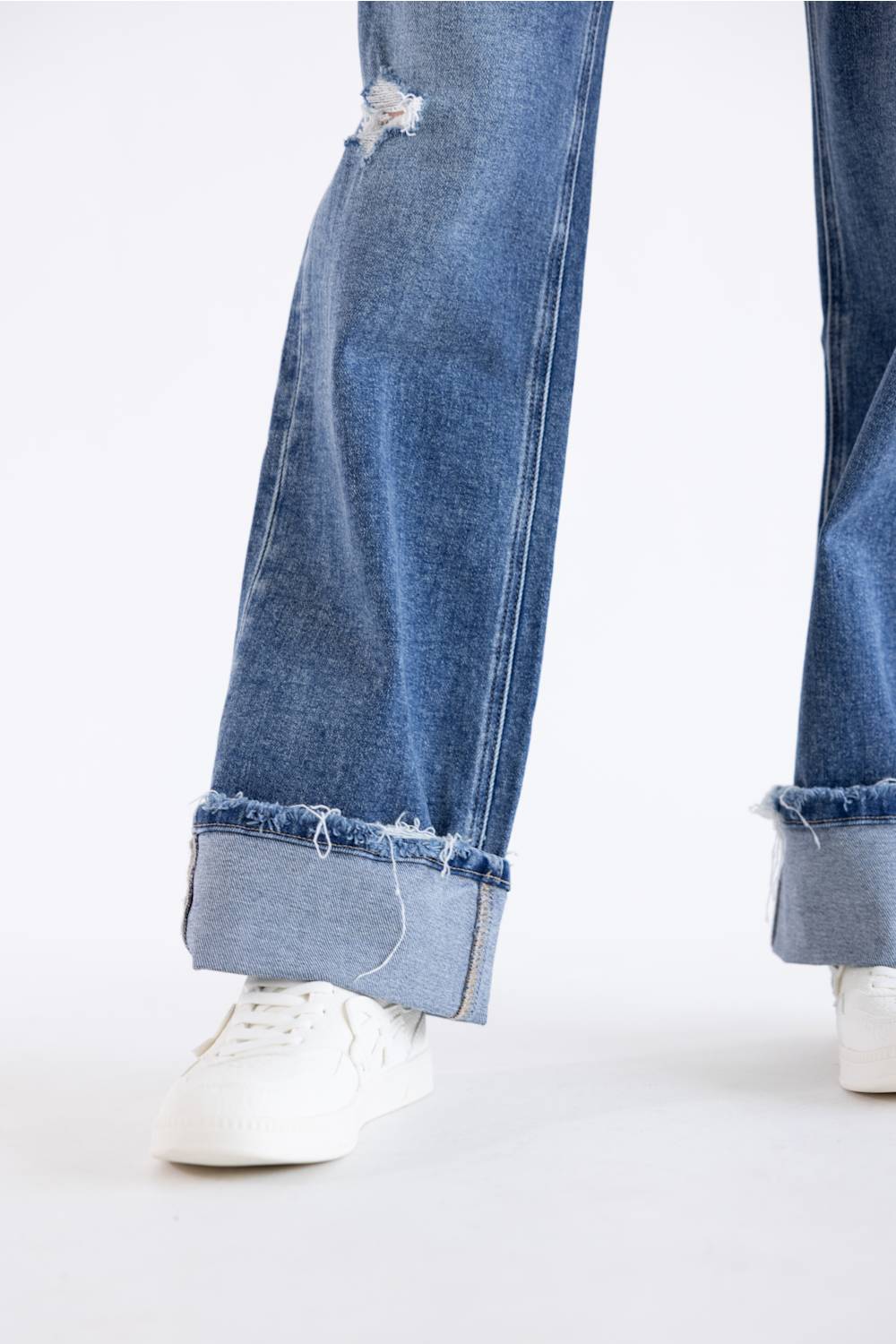 Close-up of a person's legs wearing blue wide-leg denim jeans with rolled cuffs and distressed details, paired with white sneakers on a white studio background.