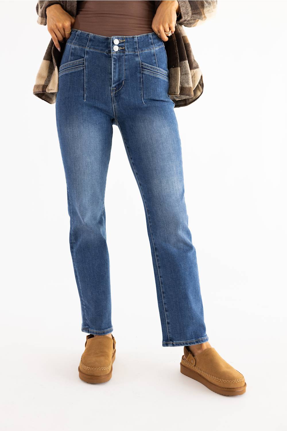 A person wearing medium-wash blue denim jeans with a brown top, a plaid shirt draped off the shoulders, and tan slip-on shoes, photographed against a plain white studio background.