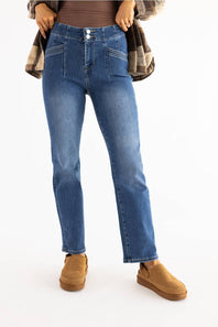 A person wearing medium-wash blue denim jeans with a brown top, a plaid shirt draped off the shoulders, and tan slip-on shoes, photographed against a plain white studio background.