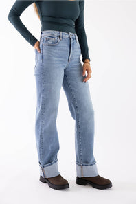A model wearing light-wash blue straight-leg jeans with rolled cuffs, paired with a dark teal long-sleeve top and brown boots, shown against a plain white studio background.