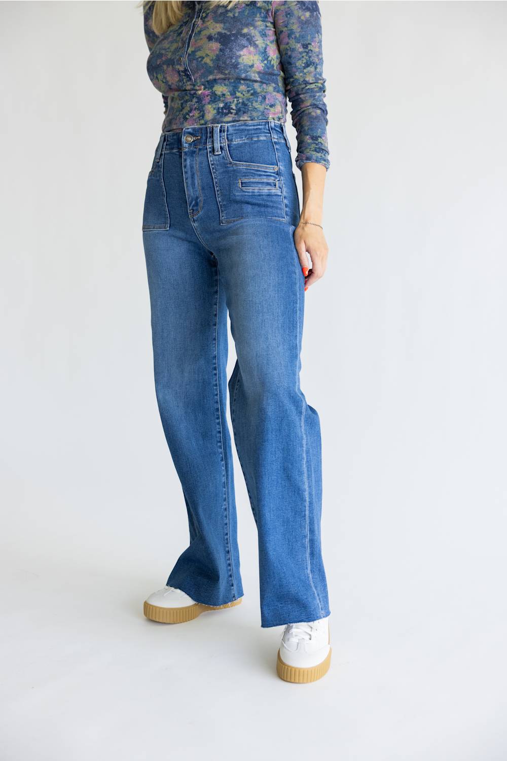 A model wearing high-waisted, wide-leg blue denim jeans with a floral long-sleeve top and white platform sneakers, photographed against a clean white studio background.
