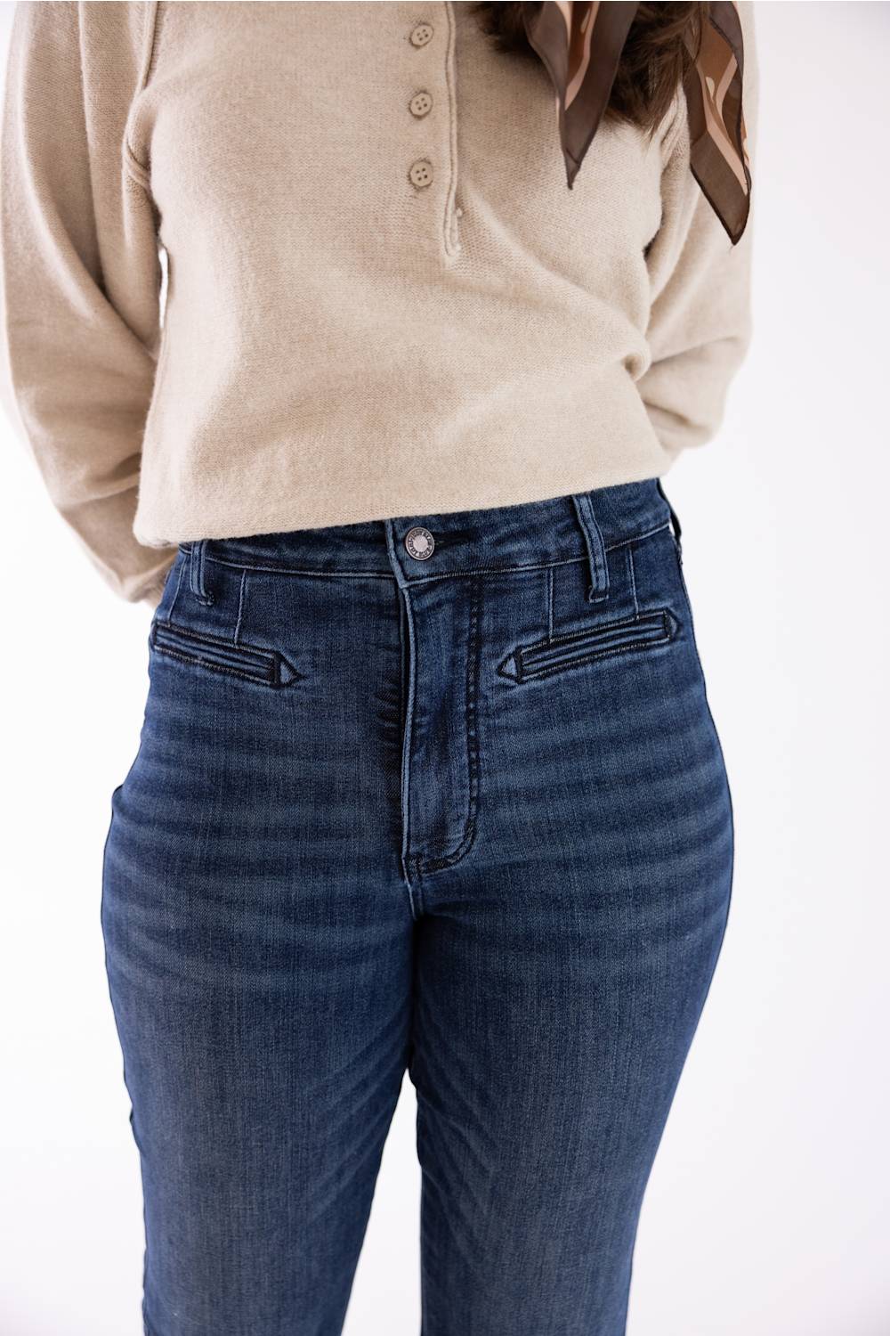 A person wearing dark blue high-rise skinny jeans and a beige henley sweater, shown from the chest down against a plain white background.