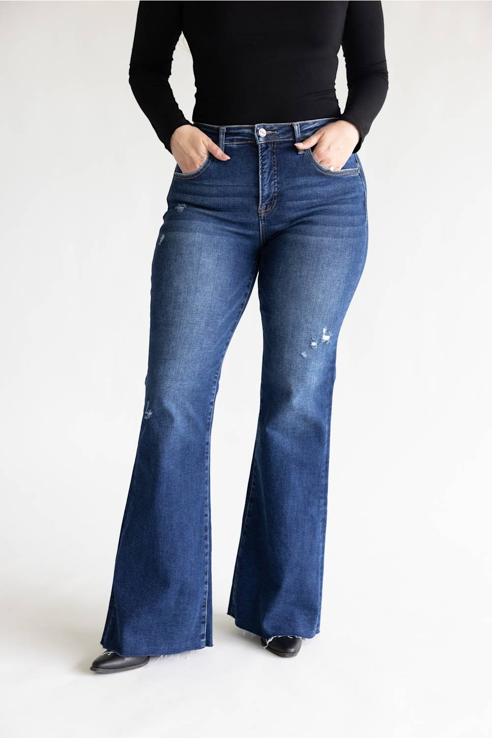 A person wearing dark blue high-waisted flare jeans with distressed details on the thighs, paired with a black long-sleeve top and black shoes, standing against a plain white studio background.